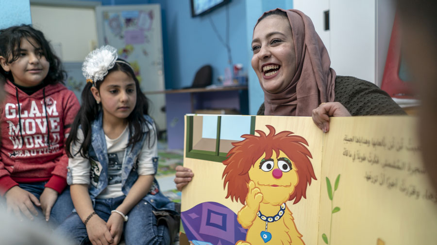 A smiling woman with a headscarf reads a book to two young girls