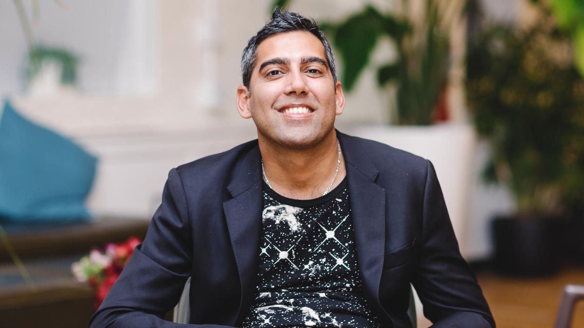 A person with short dark hair, wearing a black blazer and a black printed shirt, sits indoors in front of green plants and soft furnishings, smiling at the camera.