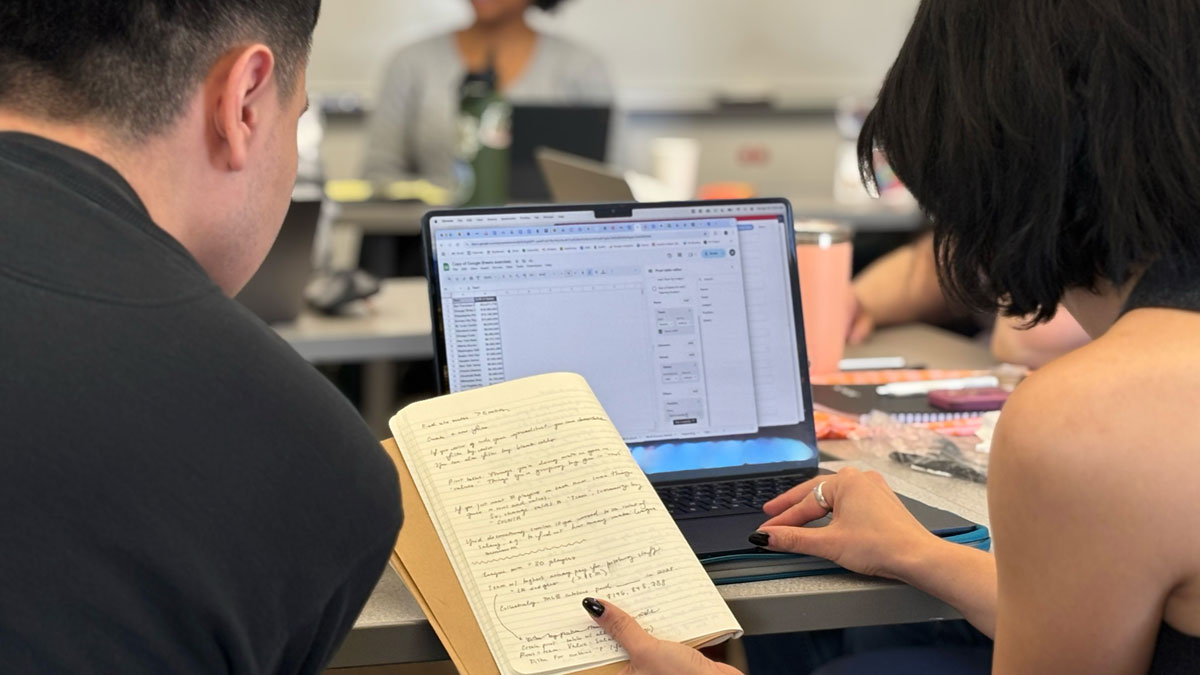 Two people collaborating with a laptop and a handwritten notebook in a classroom setting.