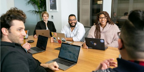 Five people sit around a table with laptops, smiling and talking during a meeting in a modern office setting.