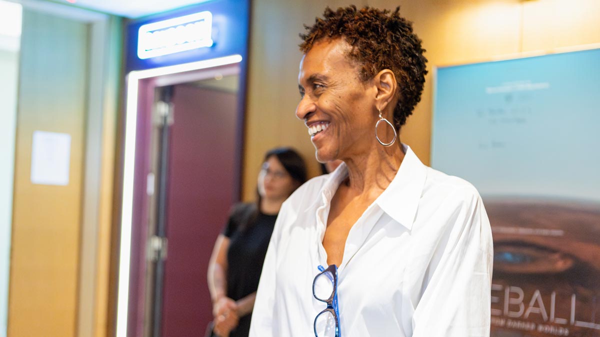 A smiling person with short curly hair in a white shirt, standing indoors near a doorway.