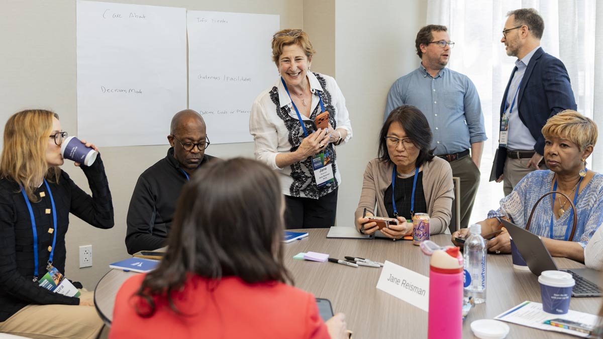 A diverse group of professionals attends a meeting around a table with a smiling woman standing near a whiteboard listing notes.