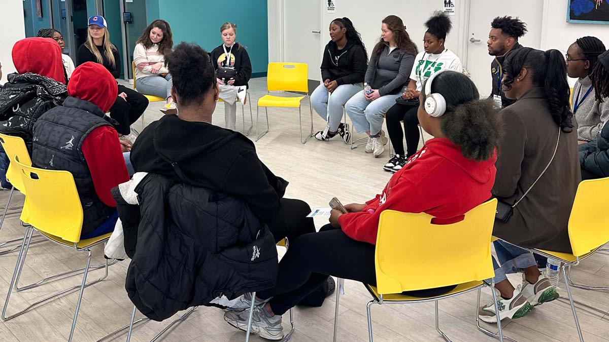 People seated in a circle on yellow chairs in a room with teal walls.