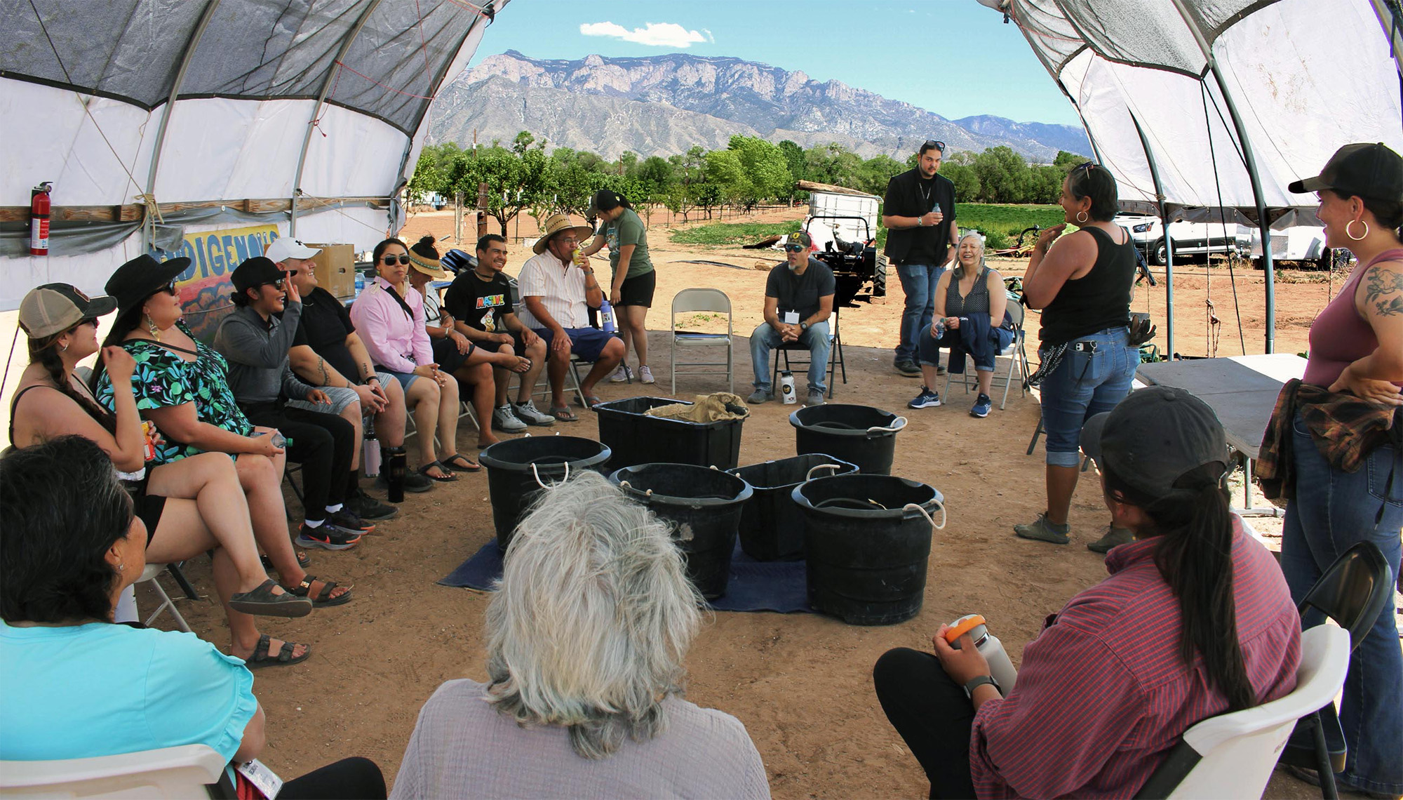 A group of people sit in a semi-circle under a tent with mountains in the background.