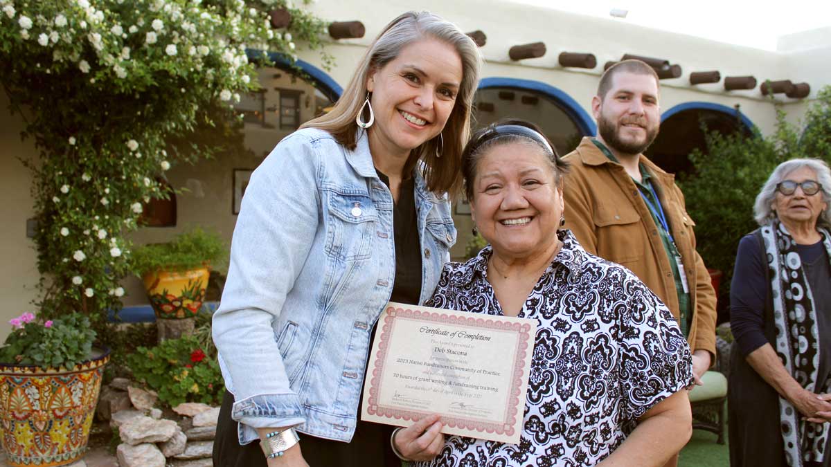 Four people outdoors, one holds a certificate, in a garden with flowers and arched windows.