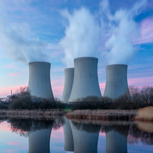 A nuclear power plant with cooling towers emitting steam, set against a colorful dawn or dusk sky and reflecting in a calm body of water.