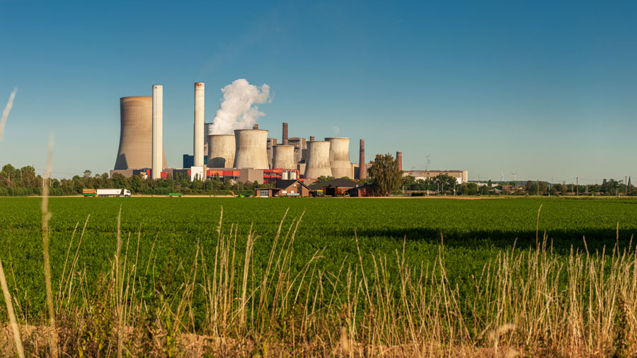 green farm field with a nuclear power plant in the distance against a blue sky