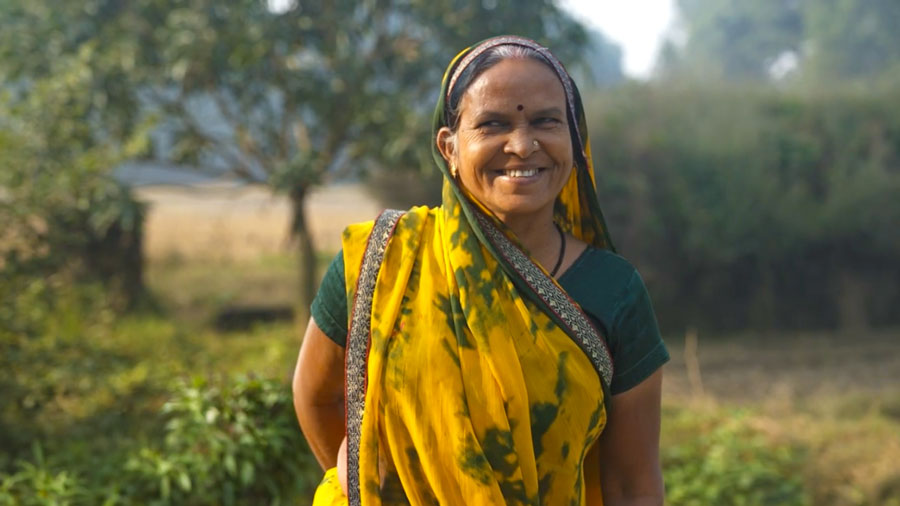 A smiling woman in a yellow and green sari stands outdoors in a rural setting.