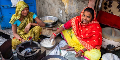 Two women in colorful saris sit on the ground preparing food, smiling near cooking utensils and bowls.