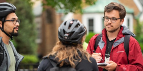 Three people, two wearing bike helmets, are having a conversation outdoors; one person is writing on a notepad.