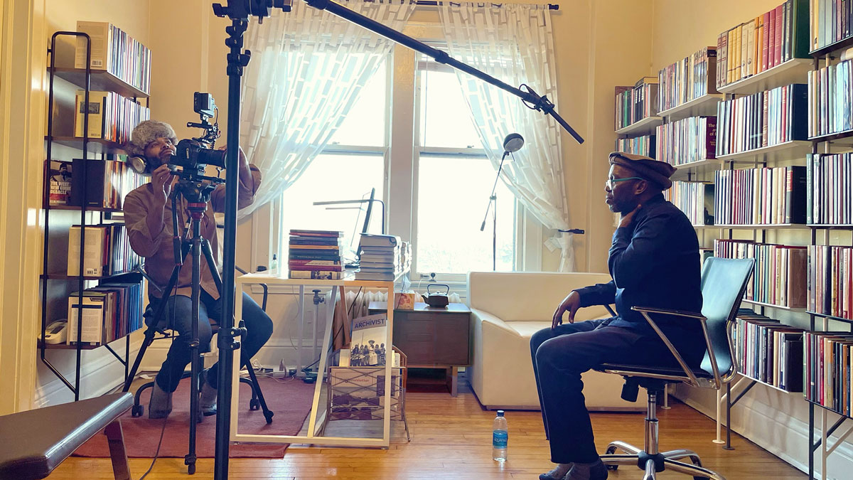 A filming session in a book-lined room with a person operating a camera and another sitting in a chair, with natural light streaming through large windows.