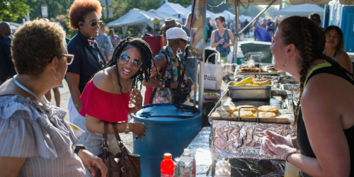 Women smile and chat at an outdoor food stall during a sunny festival with many people and tents in the background.