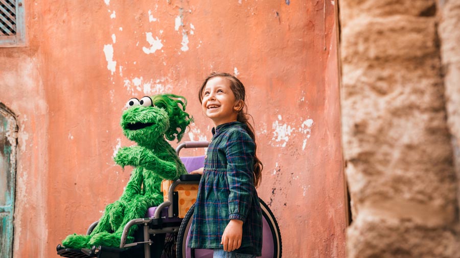 a young girl standing next to a green muppet sitting in a wheelchair