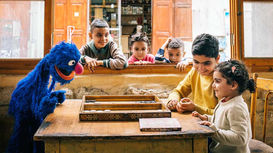 two young children play backgammon with a muppet, while three other children watch them through an open window