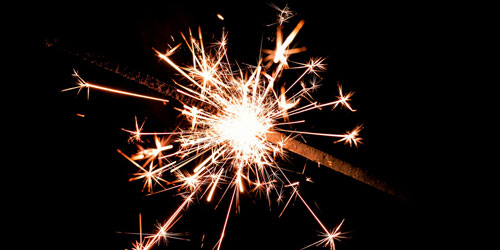 Close-up of a lit sparkler with orange and white sparks against a black background.