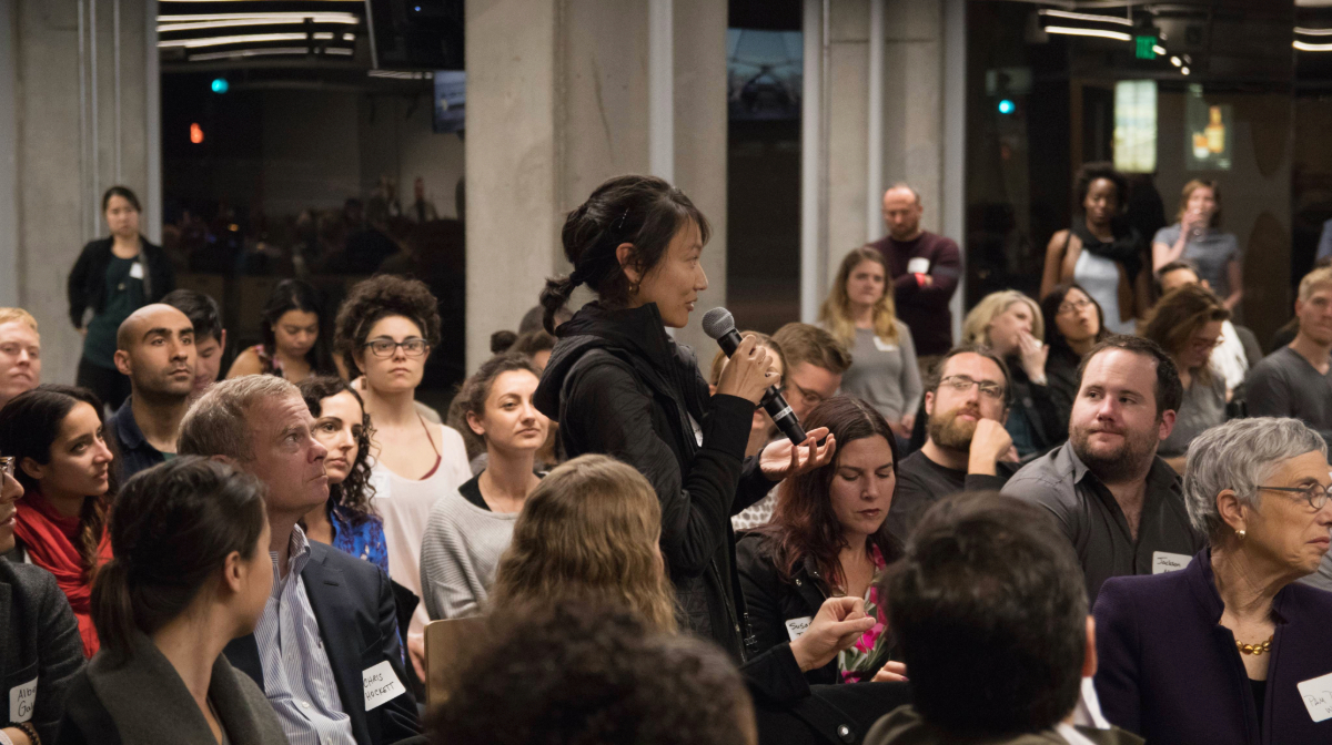 A woman stands and speaks into a microphone in a crowded room of seated and attentive people.