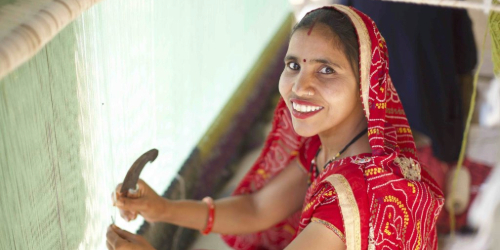 Smiling woman in a red sari weaving fabric on a loom, looking up at the camera.