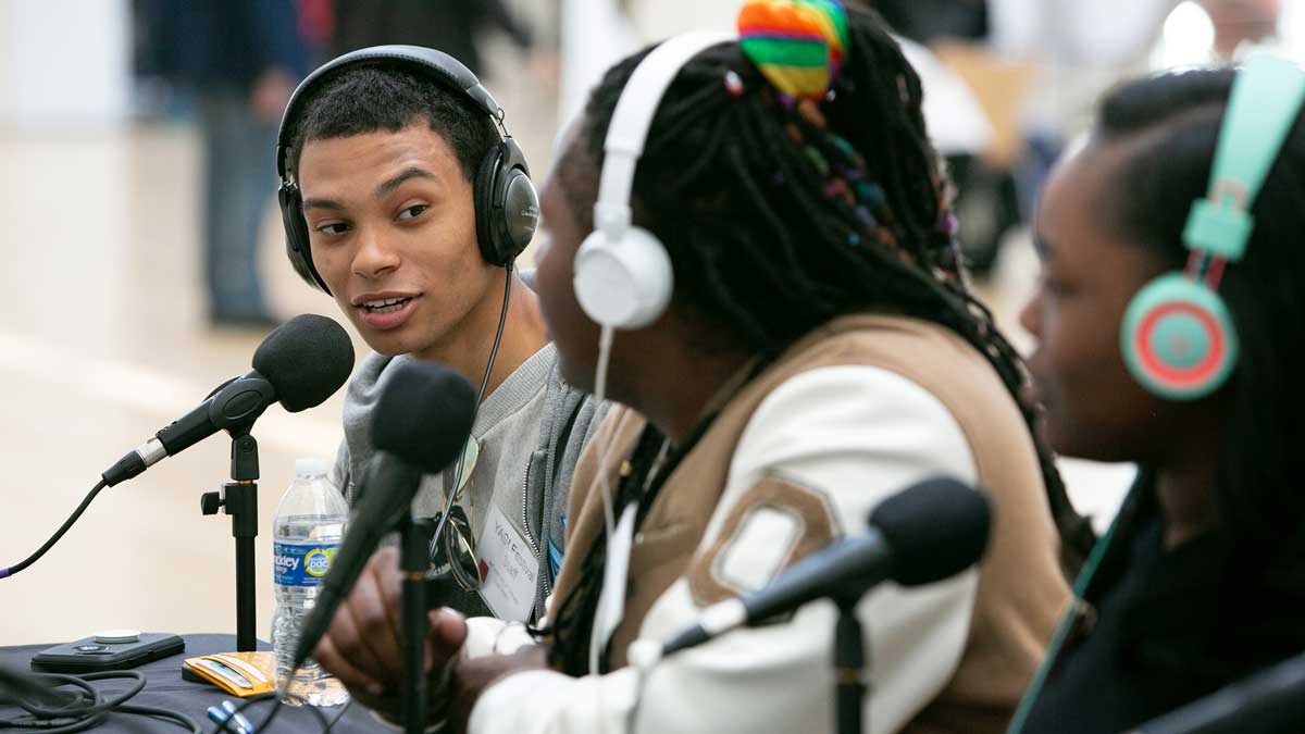 Three people wearing headphones speak into microphones at a table.