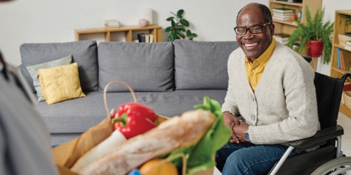 Older person in a wheelchair smiling at someone holding a grocery bag in a living room.