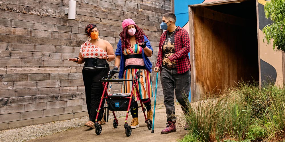 Three masked disabled people of color chat while strolling through an outdoor courtyard. On the left, an Indigenous woman wears glasses, wrist support braces, and waist trainers paired with a off-shoulder top and wide leg pants. In the middle, a Black non-binary woman walks with a rollator and wears waist trainers with ice pack inserts over their rainbow striped dress. On the right, a Latinx non-binary person walks while using a teal functional grip cane.