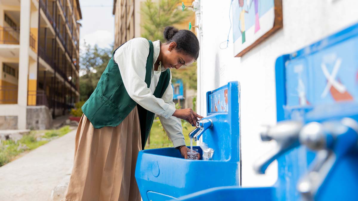 A woman in a green vest fills a cup with water at an outdoor blue water station next to a building.
