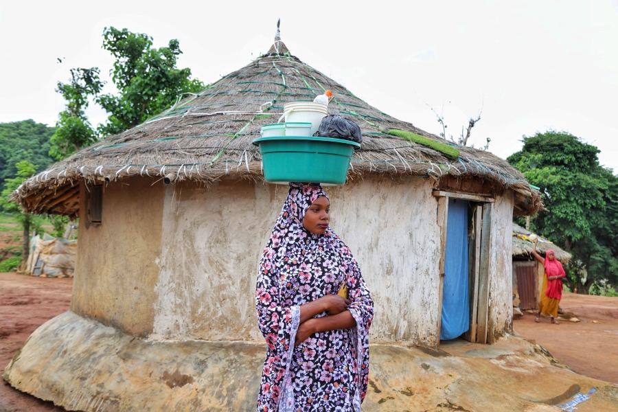 A woman standing with a bucket with supplies on her head in front of a hut.