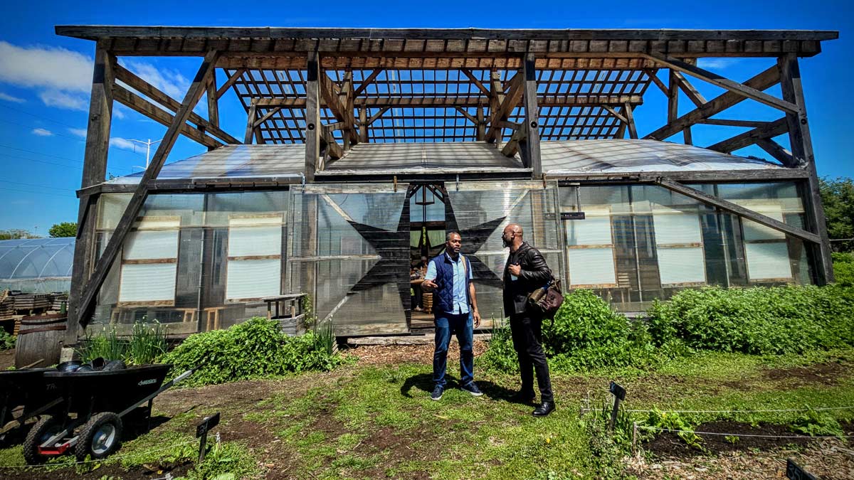 Two men stand in earnest conversation outside a vibrant community greenhouse.
