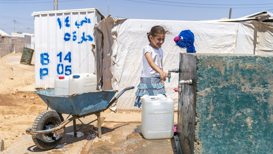 A young girl pours water into a container from a faucet while a blue Muppet standing in front of her makes her smile.