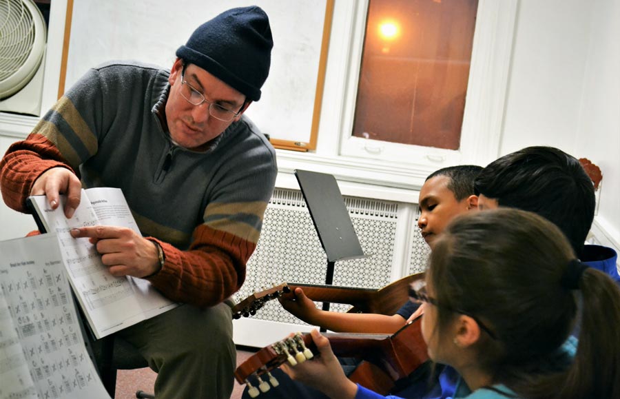 A man with a beanie hat reads a music sheet to a group of young students.