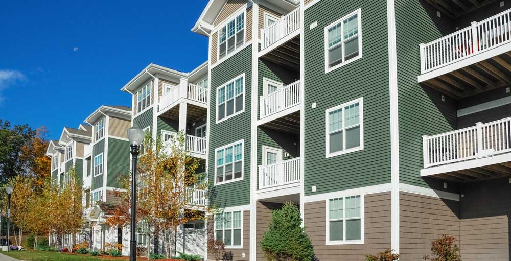 Modern apartment complex with green siding, white trim, balconies, and surrounding trees under a blue sky.