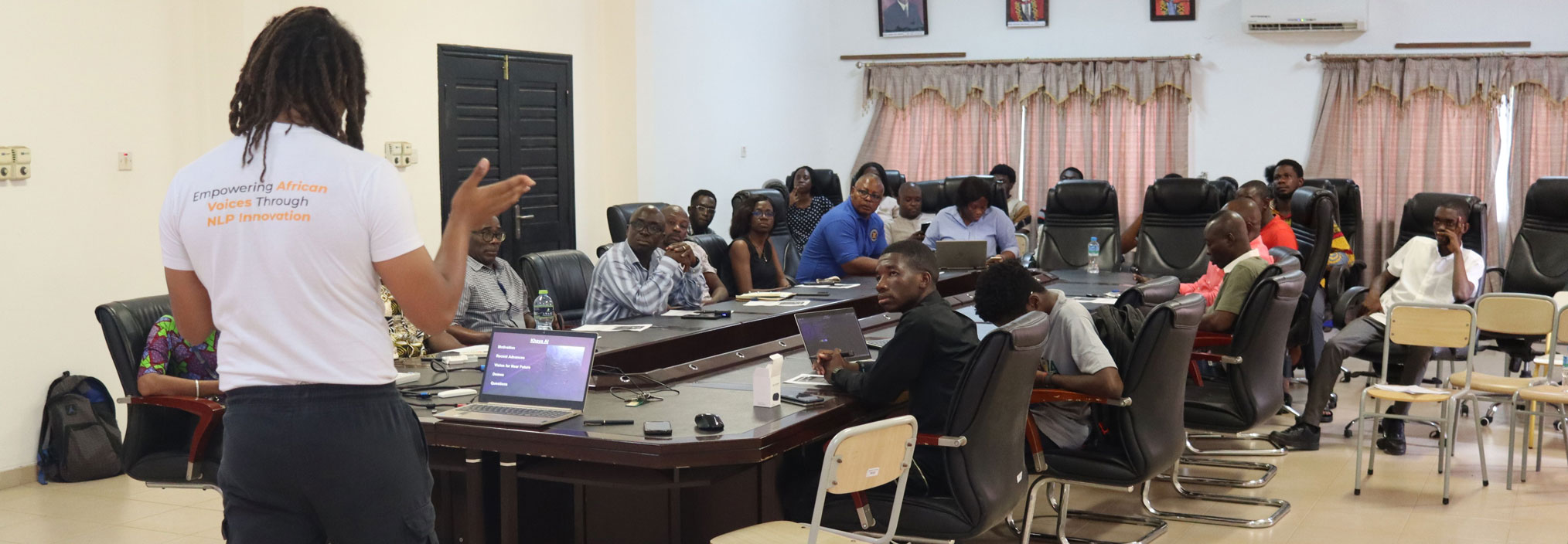 A teacher stands with his back to the camera, instructing a boardroom filled with attentive students. The teacher gestures emphatically, while referencing a laptop screen that says “OpenAI”.