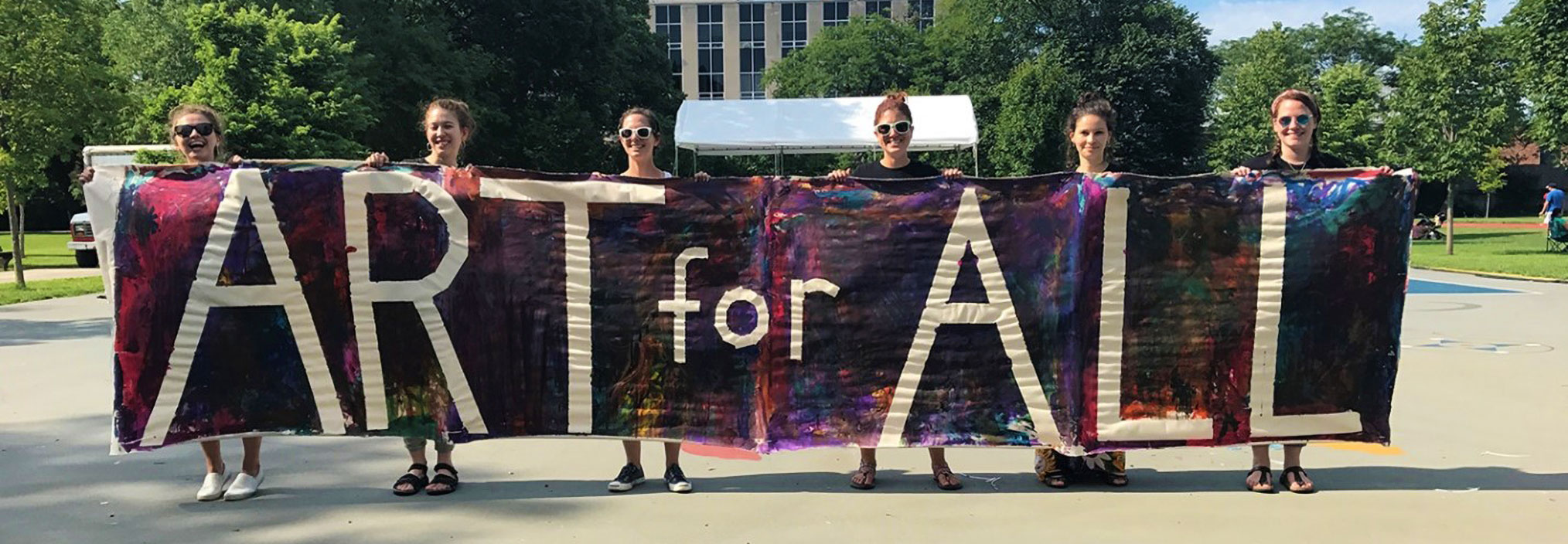 Six people holding a colorful banner with the text 'ART for ALL' in a park setting.