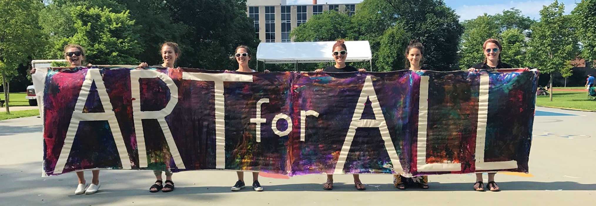 Six people holding a colorful banner with the text 'ART for ALL' in a park setting.