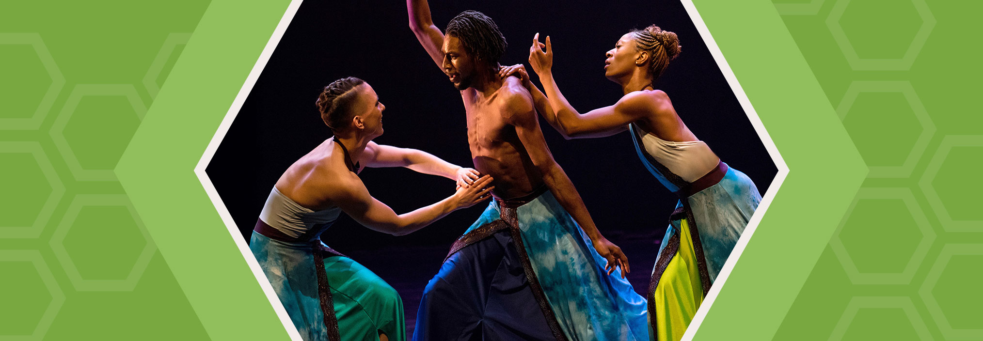Three dancers in colorful flowing costumes perform around a shirtless central dancer on a dark, stage-lit background.