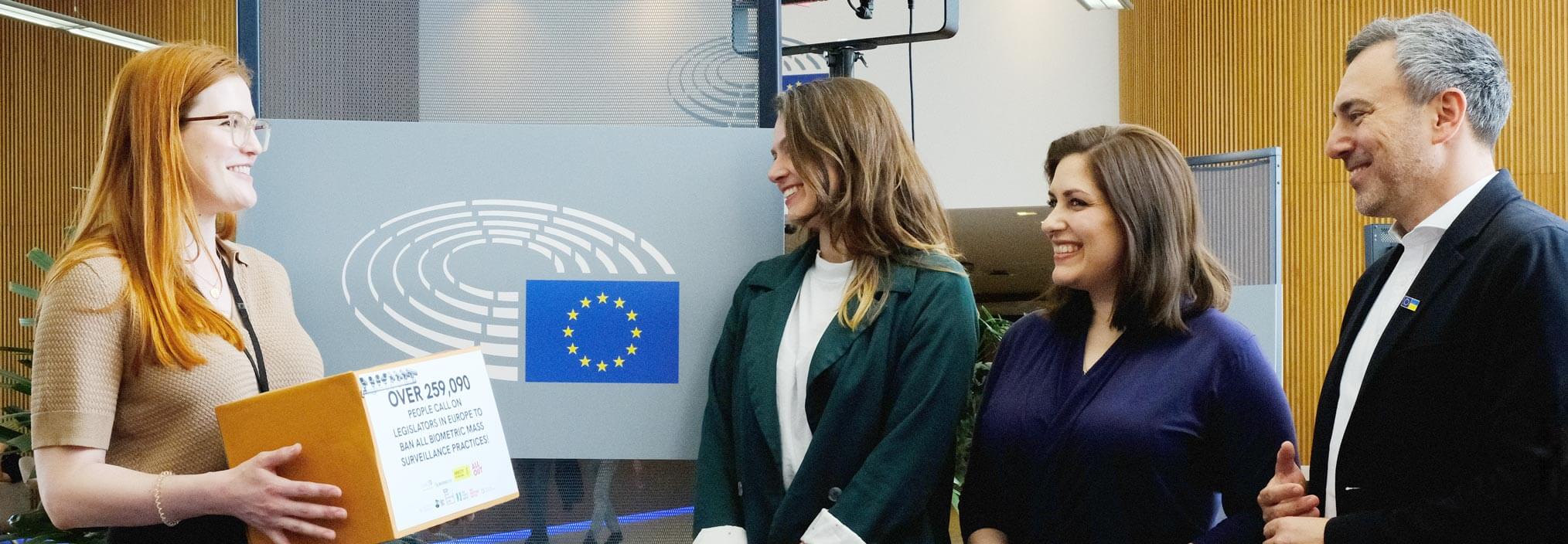 Four people stand in a formal setting, with one holding a document box. The European Union flag and Parliament logo are in the background.