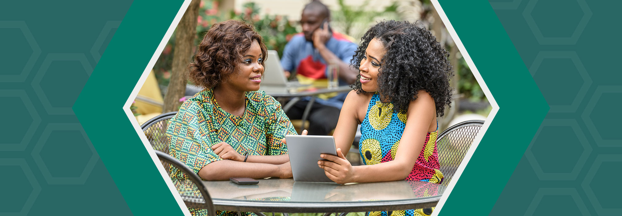 Two women seated at an outdoor table, talking and using a tablet.