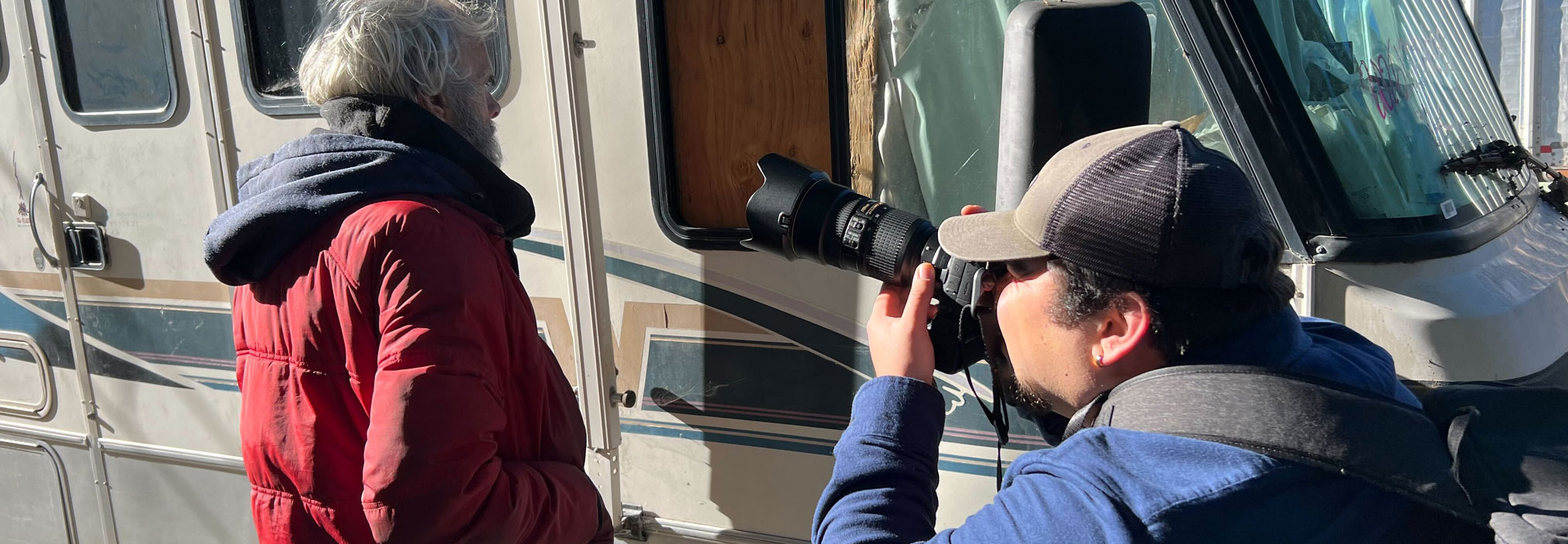 Two people stand beside a recreational vehicle while one photographs the other with a long-lens camera.
