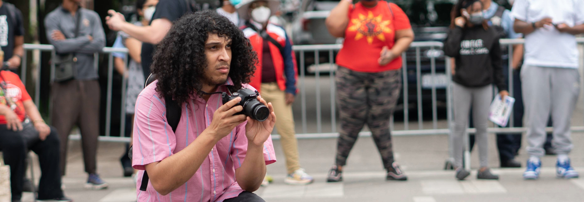 A man with curly hair and a pink shirt kneels while holding a camera at an outdoor event with people in the background.