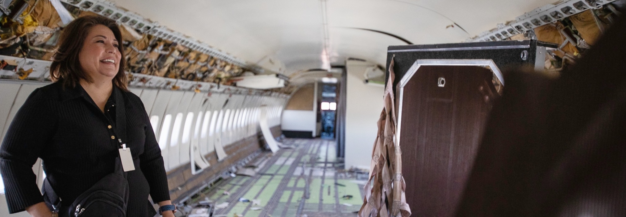 A woman smiles while standing inside an empty, stripped-down airplane cabin with exposed walls and ceiling.