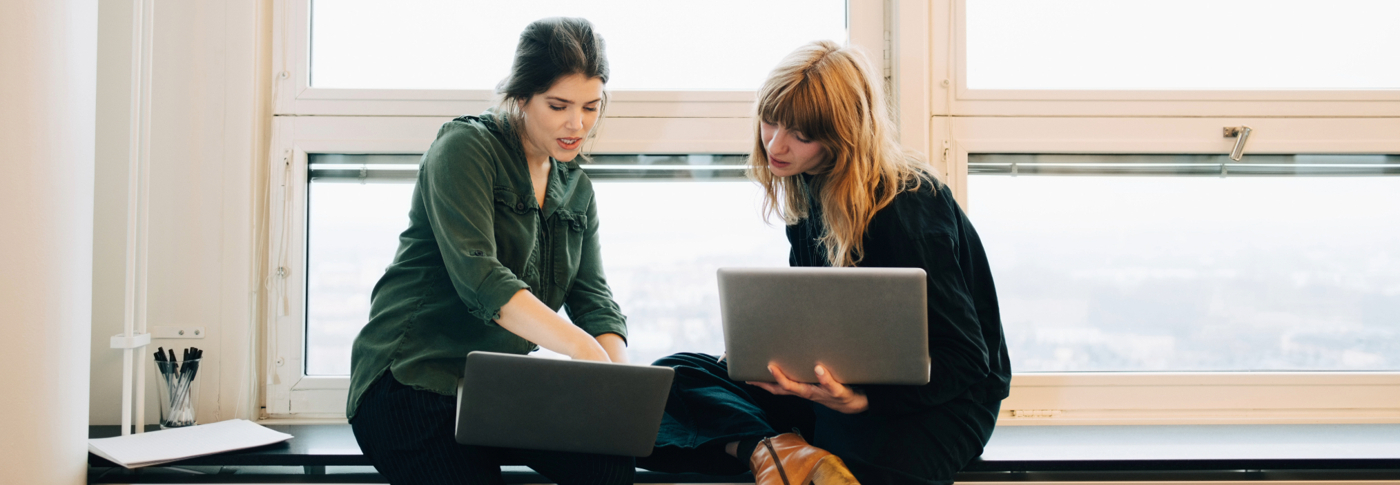 Two women sitting by a window, working together on laptops and discussing something on the screen.