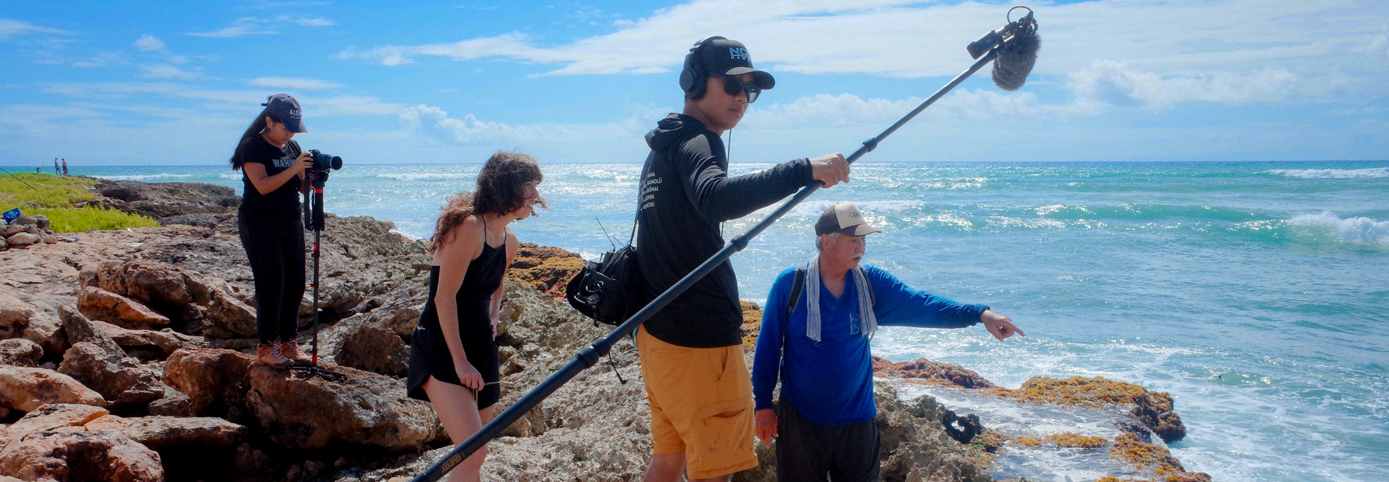 A group of people working with camera and sound equipment on a rocky shoreline by the ocean.