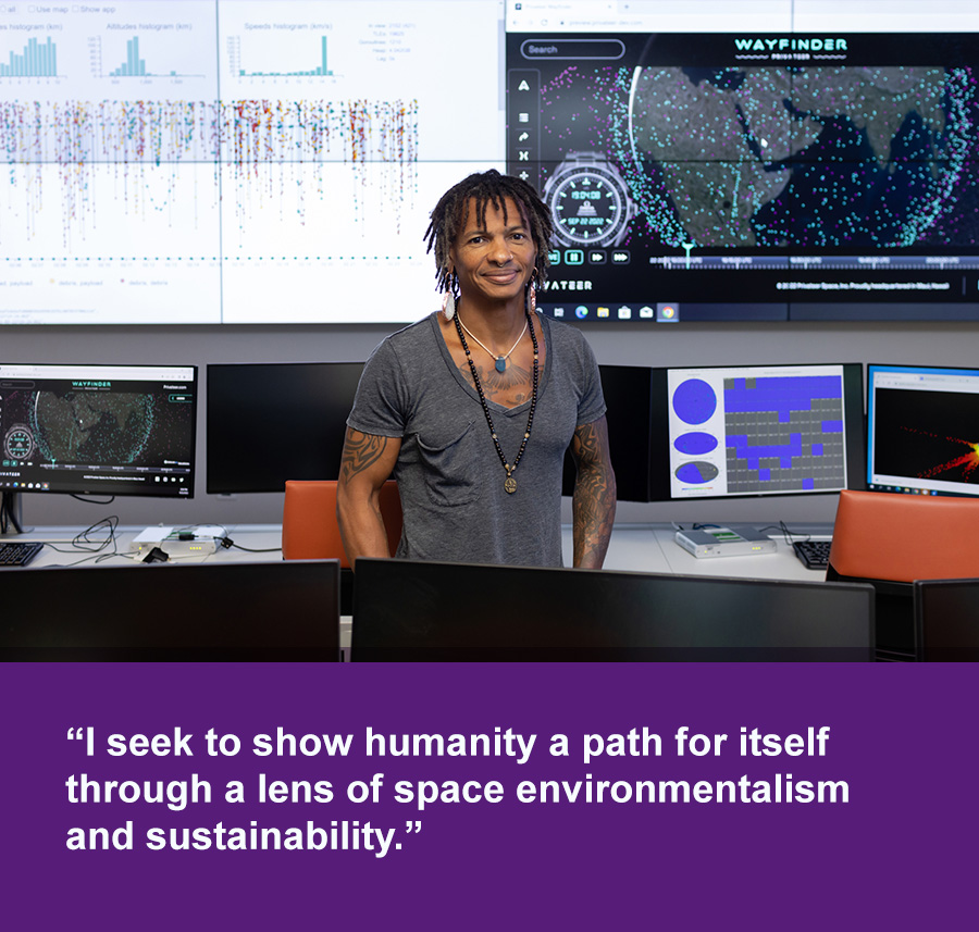 A smiling Black man stands in front of large screens with data and a digital map of the world