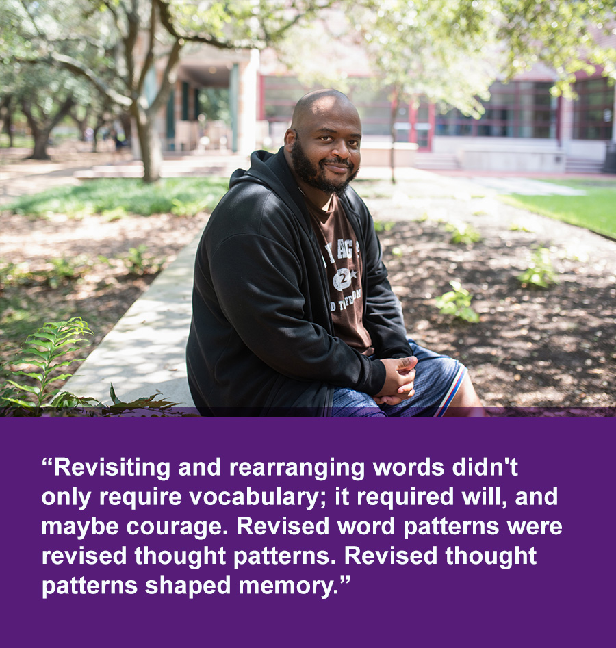 A smiling black man wearing a jacket, t-shirt, and shorts sits on a bench under shade trees on a sunny day