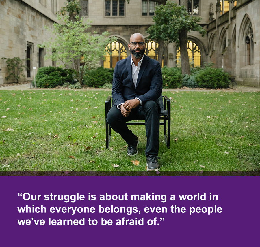 A Black man in a dark blue suit sits on a chair in a courtyard garden with a stone building in the background.