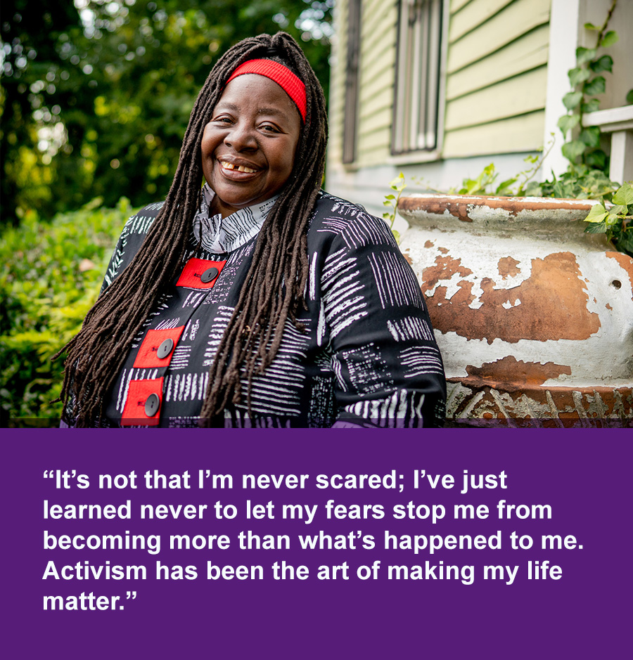 A smiling Black woman sits in front of a house with trees in the background