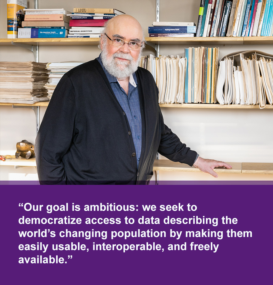 An older White man with white beard stands in front of shelves with books and files