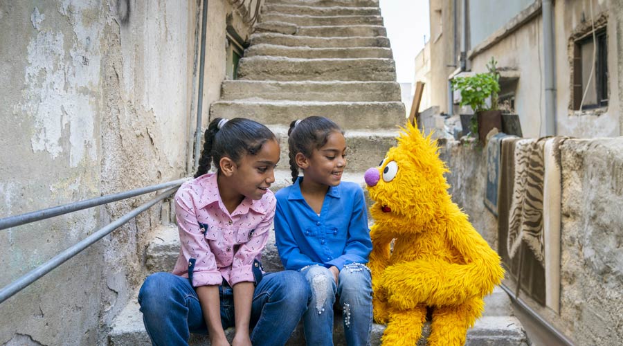 Two smiling young Syrian girls sitting on concrete stairs with yellow muppet.