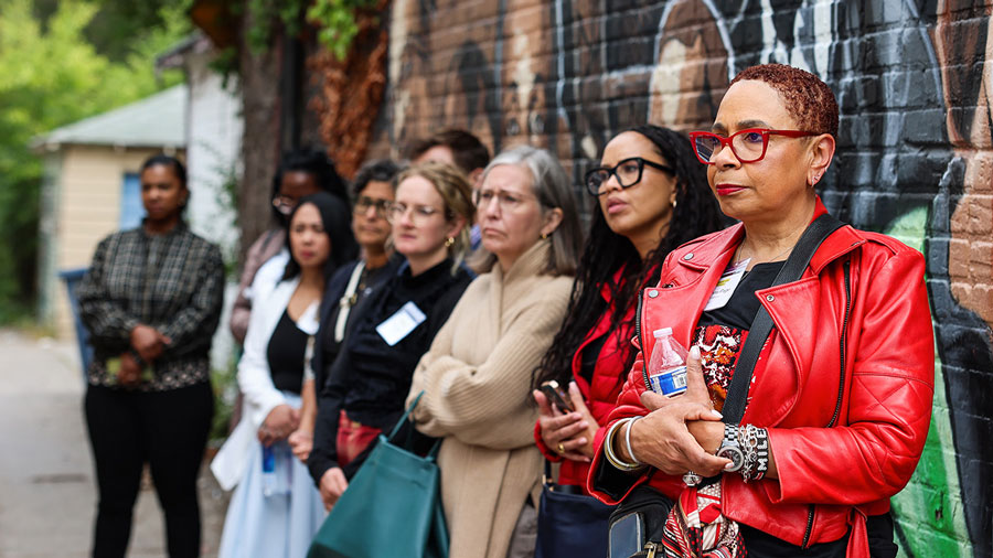 A group of women stand by a colorful mural on a brick wall in an urban alleyway, looking attentively ahead.