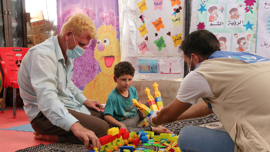 A man with white hair and a blue shirt plays with toys with a young boy while another man hands the boy more toys.