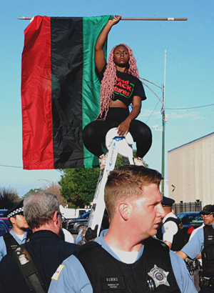 BlackWomanOnLadderHoldingUpFlagWithPoliceOfficerInForeground